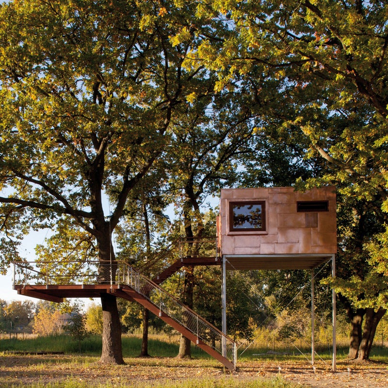 The body of this treehouse is independently supported while the terrace is suspended by cables from an oak tree and rests about a yard below the main structure. The metal of the cube was coated with a special coating to prevent oxidation and preserve the copper’s color. The interior walls and floor are dominated by pristine oiled oak paneling.