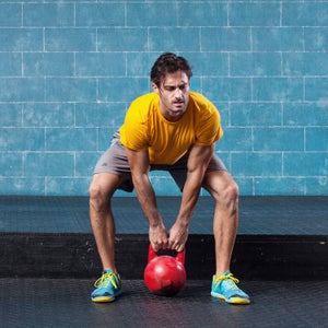 Male athlete strains at the beginning of a lift during a crossfit workout with a kettle bell.  The background is blue brick and there are several used kettle bells behind him