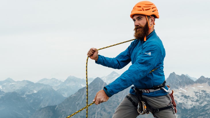 man in an orange helmet does body belay with mountains in the background