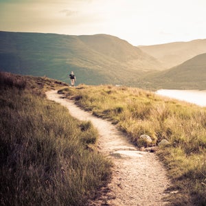 A Caucasian person is hiking through Ben Lomond by Loch Lomond, Scotland.  The person is on a dirt path with long grass on each side.  The sky is pale yellow with a few clouds.  There are dark mountains in the background and a lake in between the path and the mountains.