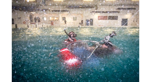 AN Tyler Sojka places a survivor into the rescue basket during a simulated rescue. The wind and the spray is provided by fans and hoses placed around the pool to mimic the intense environment under a hovering helicopter.