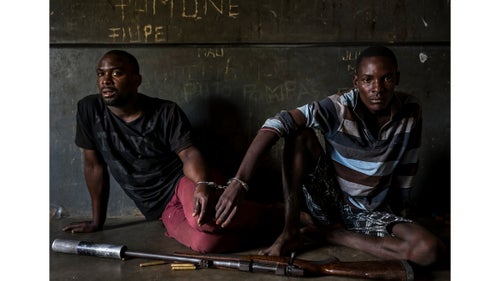 Nature - First Prize, Stories, Title: Rhino Wars
Two rhino poachers, one 19, the other 28 years old, apprehended by an anti-poaching team in Mozambique close to the Kruger National Park border. They are seen waiting to be processed in the local jail. After a three-day chase, they were caught in a roadblock and the rifle seized shortly thereafter. This was due to a coordinated effort between Kruger National Park in South Africa where the poachers intended to shoot rhino for their horns; and Sabi Game Park, a conservancy on the Mozambique side. The poachers were tracked and identified by their unique shoe pattern. They tried to say they had been on their way to buy cattle, but had no money on their persons, and, when asked, the alleged cattle owner said he did not know anything about selling his cattle. Authorities also seized a Czech CZ .458 hunting rifle, complete with a professionally built silencer. Both men admitted their guilt and will be charged under a new law which states that possession of the weapon and bullets indicates intent to poach rhino. This carries a maximum sentence of 12 years and/or $80,000. Their Toyota Hilux vehicle was also confiscated. The younger of the two poachers later led police to the homes of suspected weapons and transport suppliers, higher-ups in the rhino poaching syndicates known as level twos and threes. Those men had fled by the time the police arrived, but significant information was discovered in the form of identity documents, both real and forged, as well as banking account information.