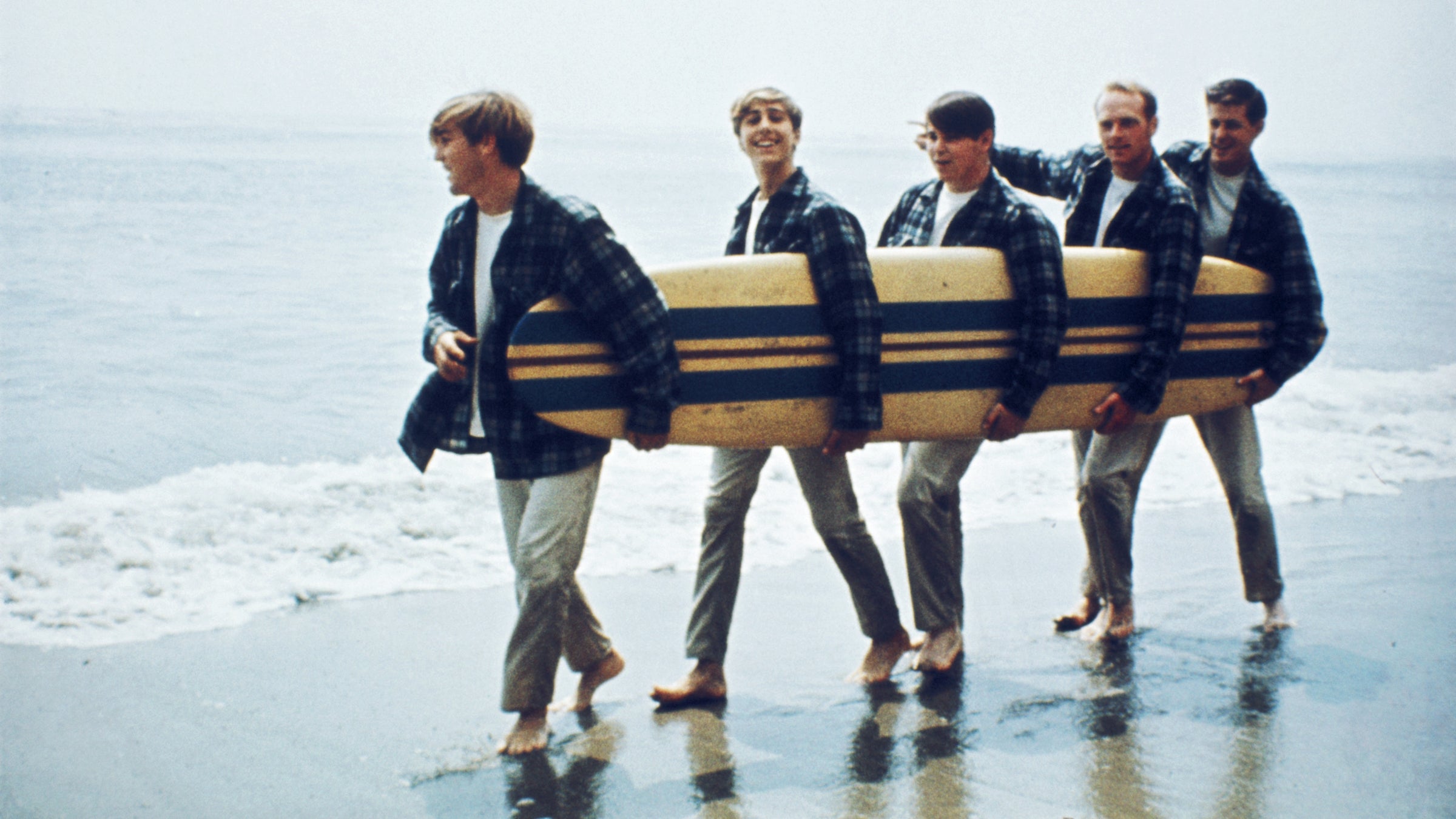 The Beach Boys walk along the beach holding a surfboard for a portrait session in August 1962