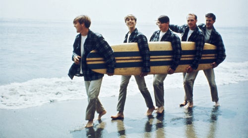 The Beach Boys walk along the beach holding a surfboard for a portrait session in August 1962