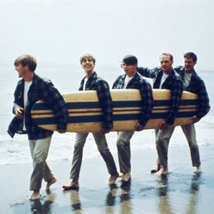The Beach Boys walk along the beach holding a surfboard for a portrait session in August 1962