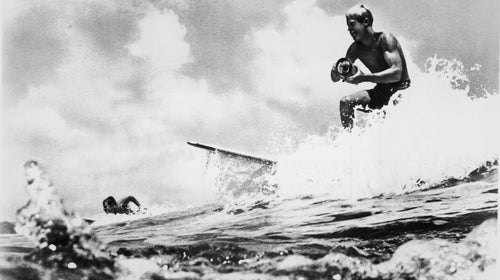 American surfer and director Bruce Brown surfs a wave and holds a camera while filming footage for his international surfing documentary, 'The Endless Summer,' circa 1966.
