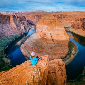 A woman sits on a cliff edge above the Horseshoe Bend of the Colorado River.