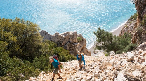 Two men hike up steep talus slope above sea, with climbing rope