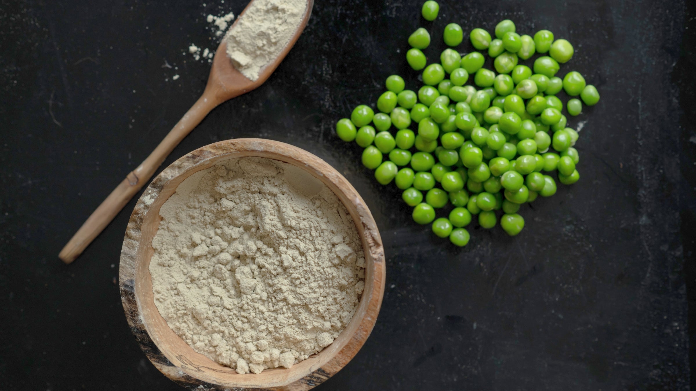 Pea protein powder is pictured in a wooden bowl, and in a wooden spoon from an aerial view. Next to them is a pile of peas.