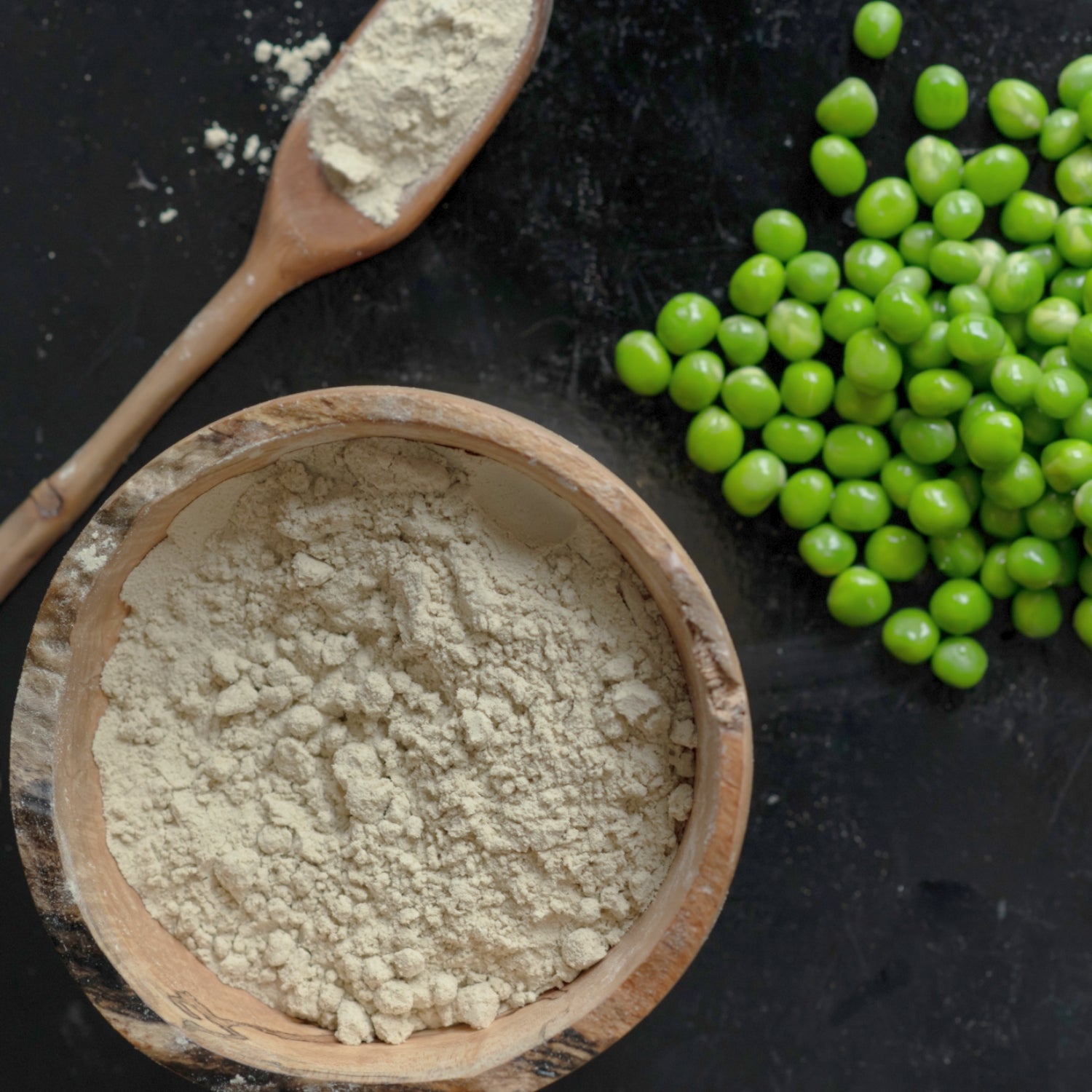 Pea protein powder is pictured in a wooden bowl, and in a wooden spoon from an aerial view. Next to them is a pile of peas.