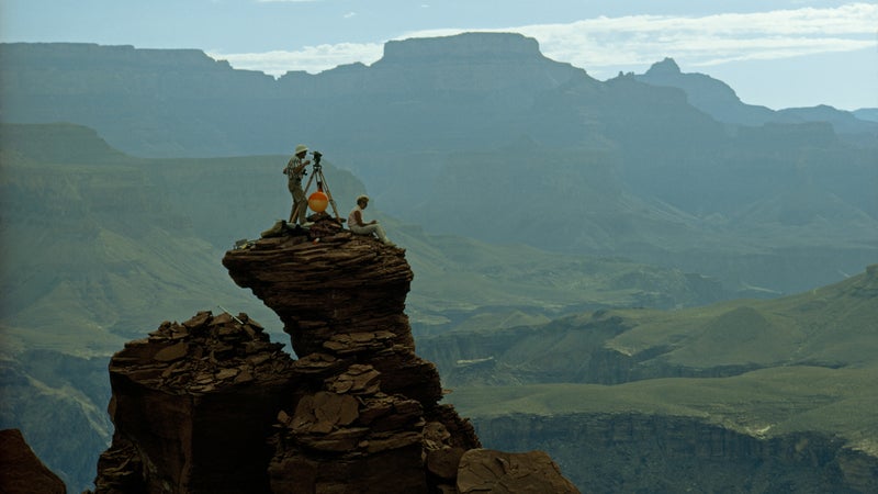 Bradford Washburn stands atop Dana Butte as he uses a surveying instrument called a theodolite to measure the horizontal and vertical angles between two points in the Grand Canyon. His wife, Barbara, sits next to him recording the data. Some of the surveying points used for the map could only be reached by helicopter.