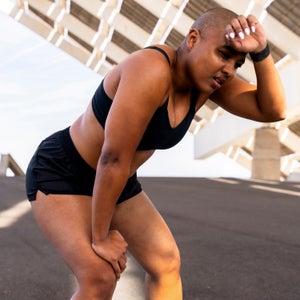 Exhausted sporty woman with hand on knee at road, sweating while training in heat