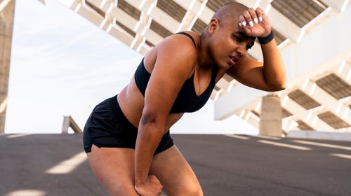 Exhausted sporty woman with hand on knee at road, sweating while training in heat