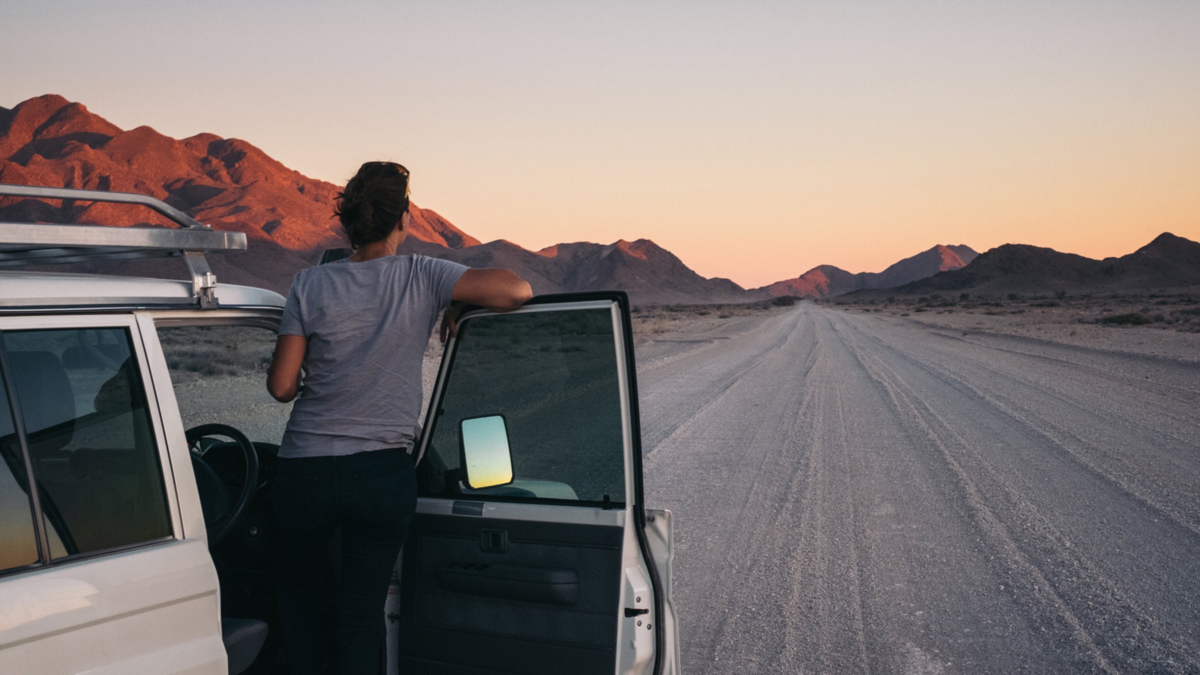 Woman On A Road Trip Through The Desert Leaning Out Of Her Car Watching A Sunset