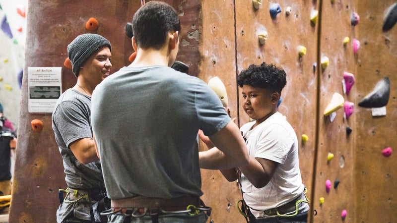 Two Brown Ascenders show a mentee how a belay device works before he climbs.