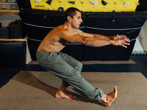 Man does a pistol squat on his left leg, which is bent at a 90-degree angle as he hovers his straightened right leg above the gym floor.
