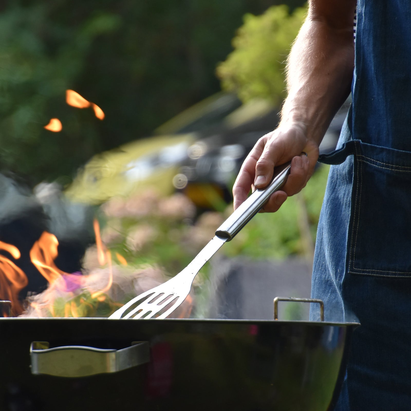 Male arm holding a spatula over a flaming grill wearing an apron