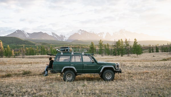 Two Anonymous Men Sitting On The Green Jeep Parked In The Wild Area Surrounded By Mountains