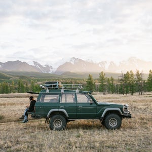 Two Anonymous Men Sitting On The Green Jeep Parked In The Wild Area Surrounded By Mountains