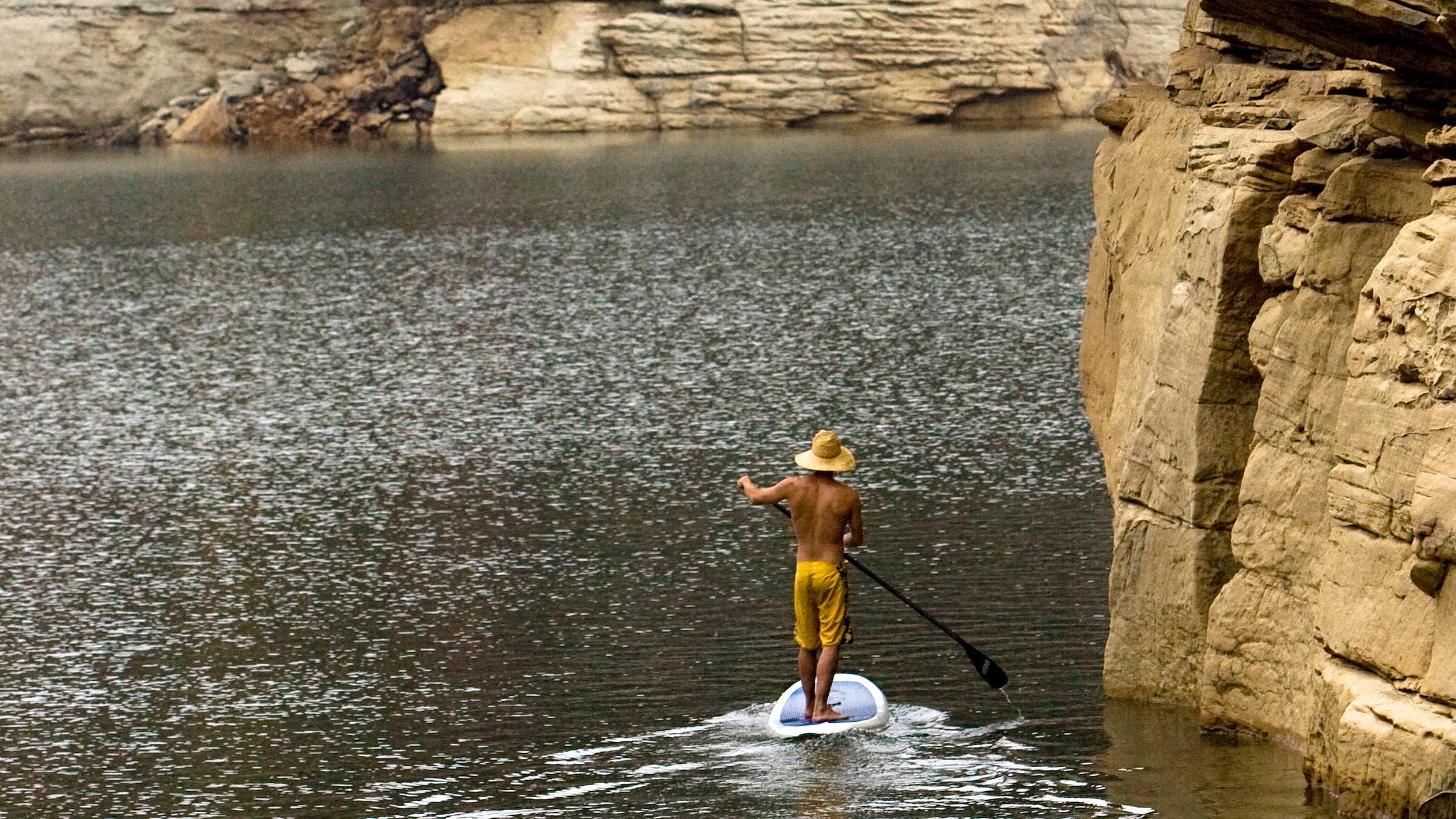 One man stand up paddleboarding on a lake under big cliffs with fall colors.