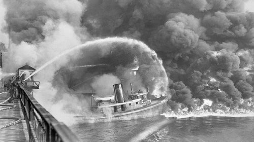 From November 3, 1952: Firemen stand on a Cleveland bridge over the Cuyahoga River, spraying water on the tug Arizona, as a fire, started in an oil slick, swept the docks at the Great Lakes Towing Company site on November 1. The blaze destroyed three tugs, three buildings, and the ship-repair yards.