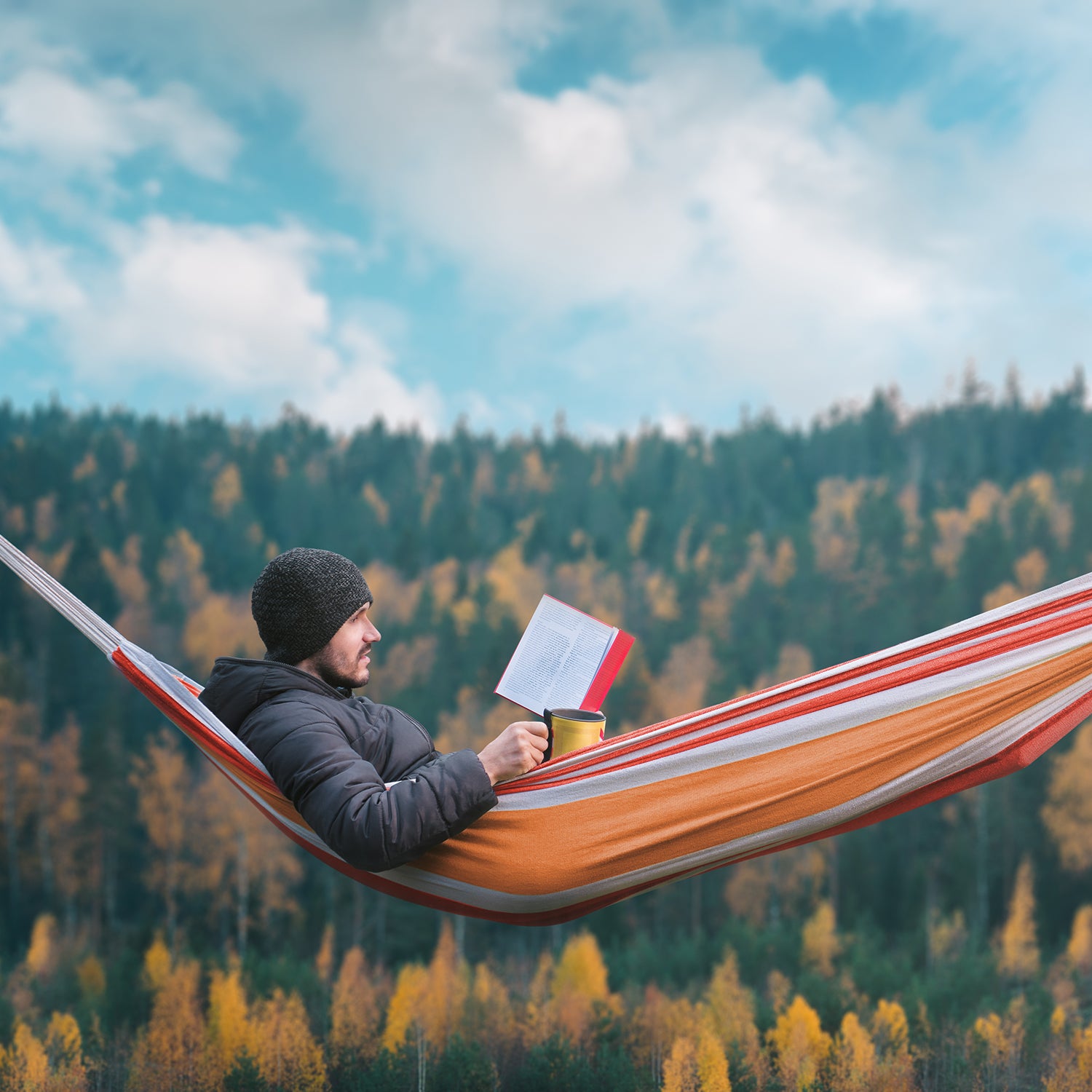 A man sits in a hammock and reads a book in a picturesque place. Mug in his right hand.