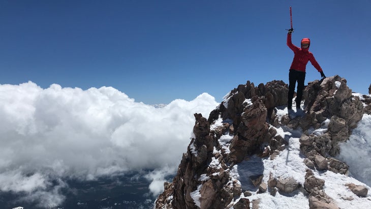 A climber in a red jacket holds an ice axe above her head in celebration at the summit of Mount Shasta