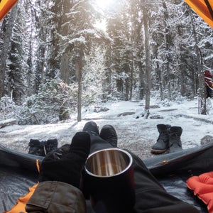 Camping with hand holding cup in yellow tent with snow in pine forest