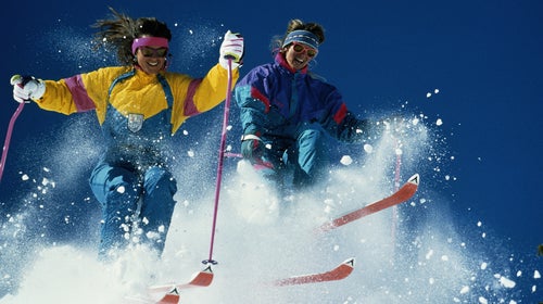 Two women skiing downhill, creating snow spray, low angle view