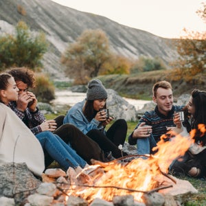 Smiling charismatic friends different races at camping, sitting beside a bonfire and drinking some hot drinks from metallic cups and spending a amazing time together.