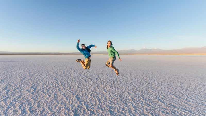Two Male Friends Jumping Happy On Salt Flat Desert Landscape While Adventure Travel