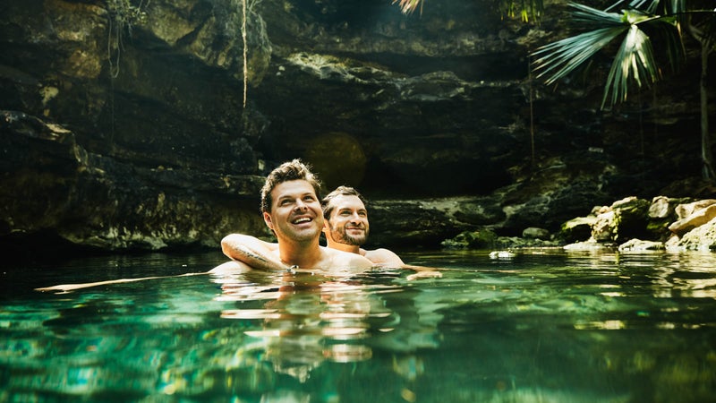Smiling gay couple embracing while swimming in cenote during vacation