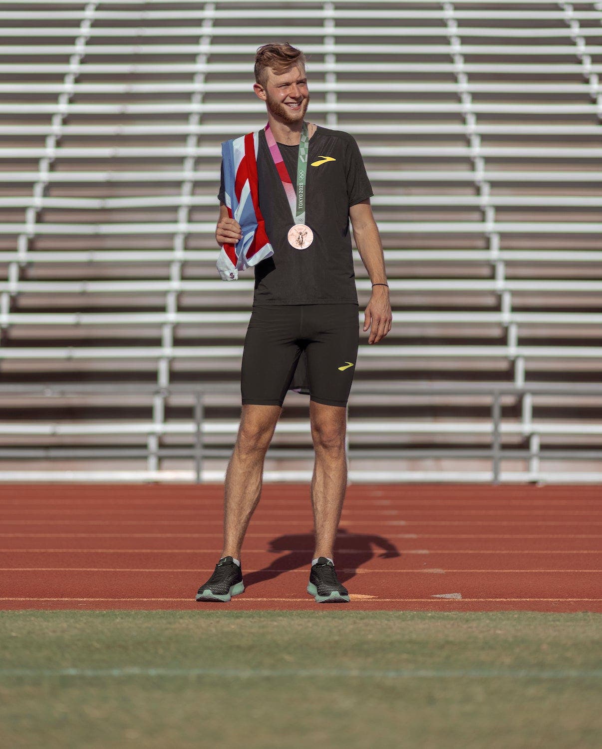 Josh Kerr standing on track with bronze medal around neck and holding Great Britain flag over his right shoulder.