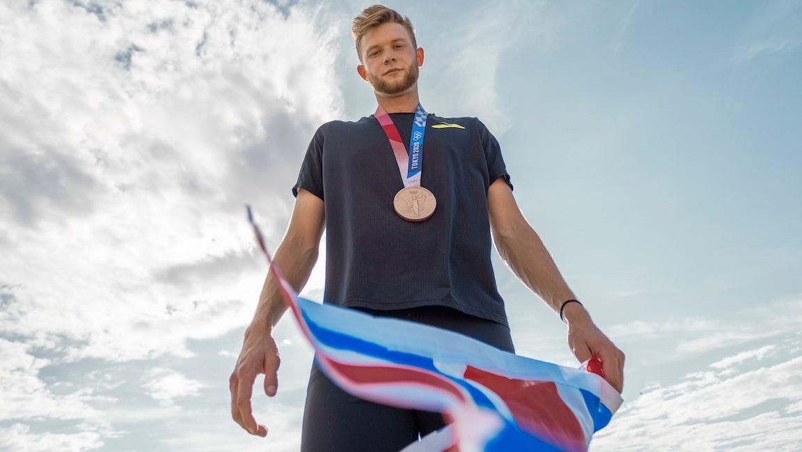 Josh Kerr with bronze medal around neck looking down on camera with Great Britain flag in hand.