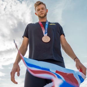 Josh Kerr with bronze medal around neck looking down on camera with Great Britain flag in hand.