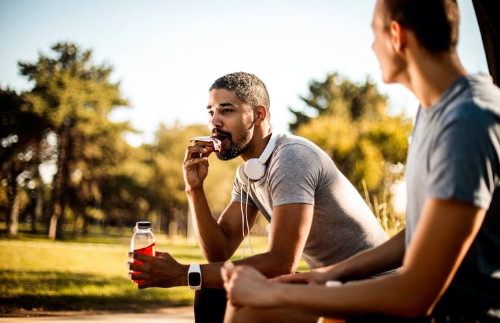 Active young man taking a bite out of an energy bar and hydrating himself with a sport drink next to his friend.