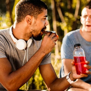 Active young man taking a bite out of an energy bar and hydrating himself with a sport drink next to his friend.