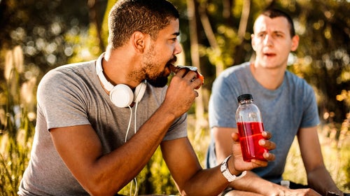 Active young man taking a bite out of an energy bar and hydrating himself with a sport drink next to his friend.