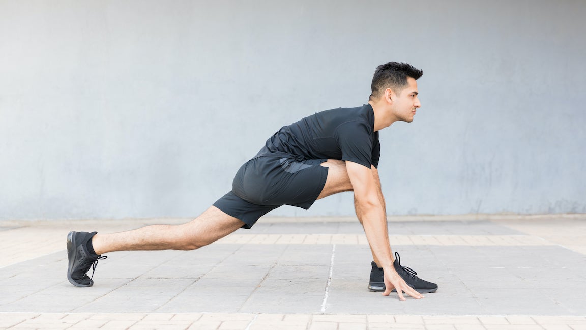 Man with black hair in black t-shirt and black shorts doing a yoga pose in which one leg is straight back on the ground and the other is on the ground with his knee bent at a 45-degree angle as he bends over into a lunge-like position.