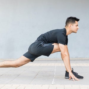 Man with black hair in black t-shirt and black shorts doing a yoga pose in which one leg is straight back on the ground and the other is on the ground with his knee bent at a 45-degree angle as he bends over into a lunge-like position.