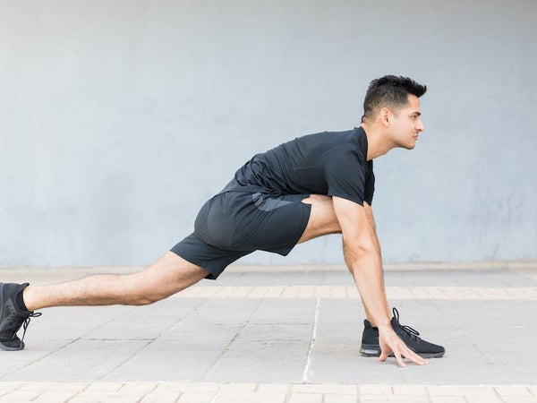 Man with black hair in black t-shirt and black shorts doing a yoga pose in which one leg is straight back on the ground and the other is on the ground with his knee bent at a 45-degree angle as he bends over into a lunge-like position.