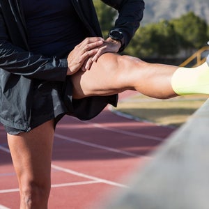 Anonymous male runner leaning on railing and stretching legs while warming up before training at stadium on sunny day.