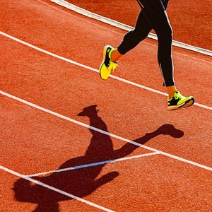 Sportsperson running over the orange running track. Sports clothing - sports shoes and running tights. High angle view of a runner's legs and its deep shadow on the track.