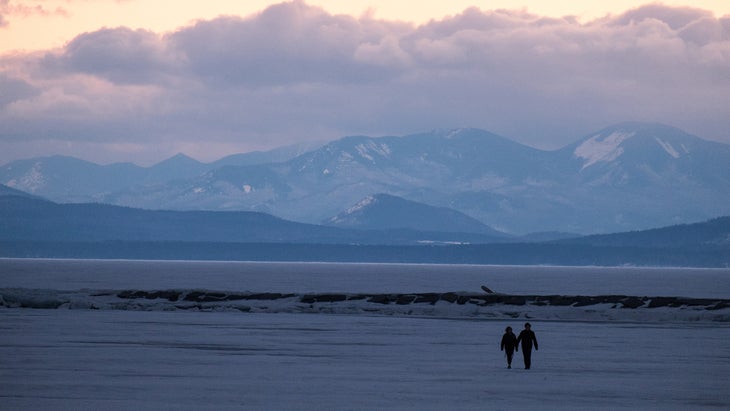 A couple walks across the ice on frozen Lake Champlain in Burlington Vermont at Sunset. We can see the snow capped mountains and New York State in the distance.