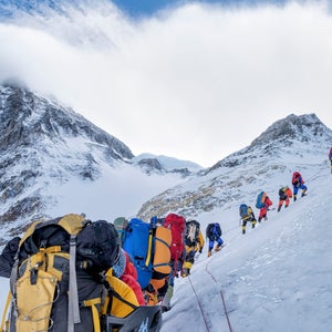 A line of climbers wearing brightly colored backpacks and coats climb up a snowy Mount Everest.