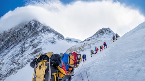 A line of climbers wearing brightly colored backpacks and coats climb up a snowy Mount Everest.