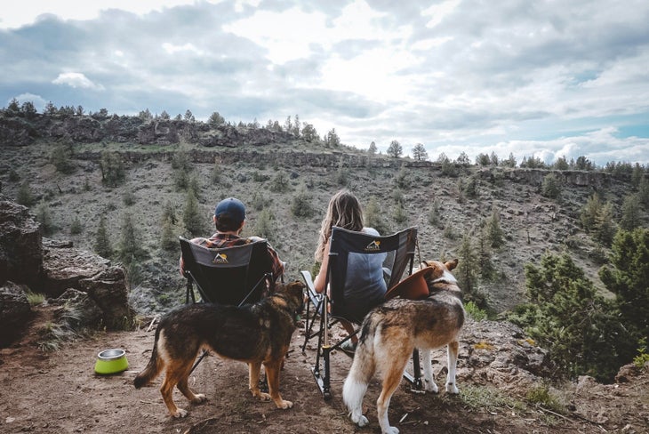 115705475_10164119931815714_3264130705653968410_o A man and a woman sitting in camp chairs looking over a tree-covered hillside with two dogs behind them.