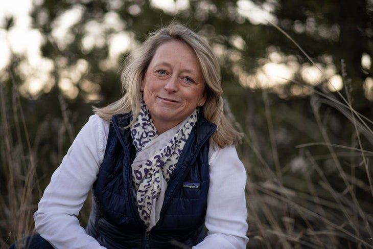 Woman with long blonde hair, white shirt, navy blue vest and scarf smiling, in field with tall grasses