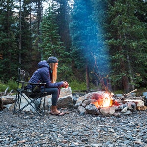 A woman sitting in a folding chair in front of a campfire in the woods