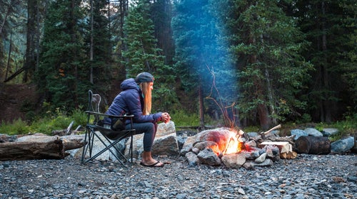 A woman sitting in a folding chair in front of a campfire in the woods